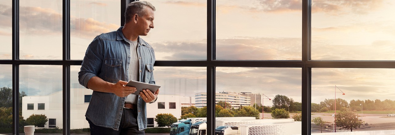 Ein Mann mit einem Tablet steht an einem Fenster und blickt auf seinen Lkw-Fuhrpark
